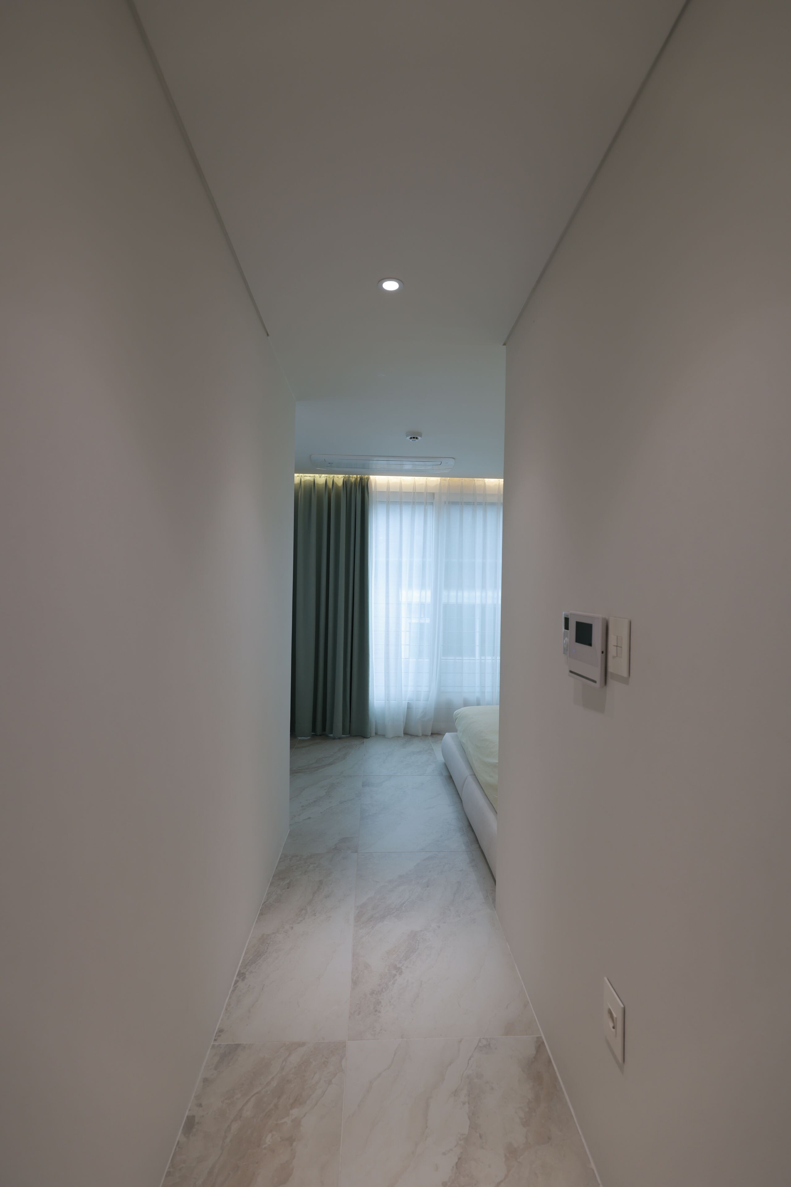 A hallway leading to a bedroom with a bed, a window with curtains, and a ceiling light.
