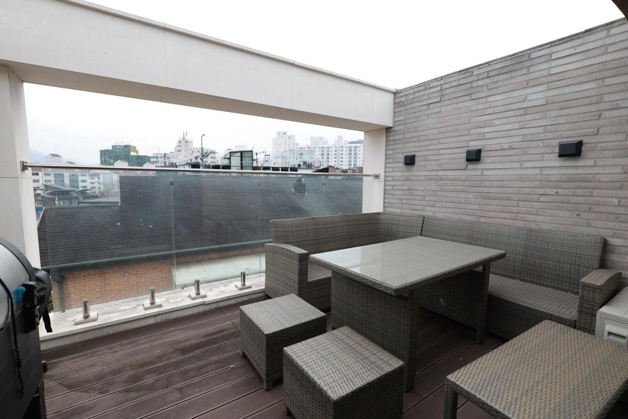 Un balcón exterior con vistas a la ciudad, amueblado con mobiliario de mimbre y una mesa de vidrio.
