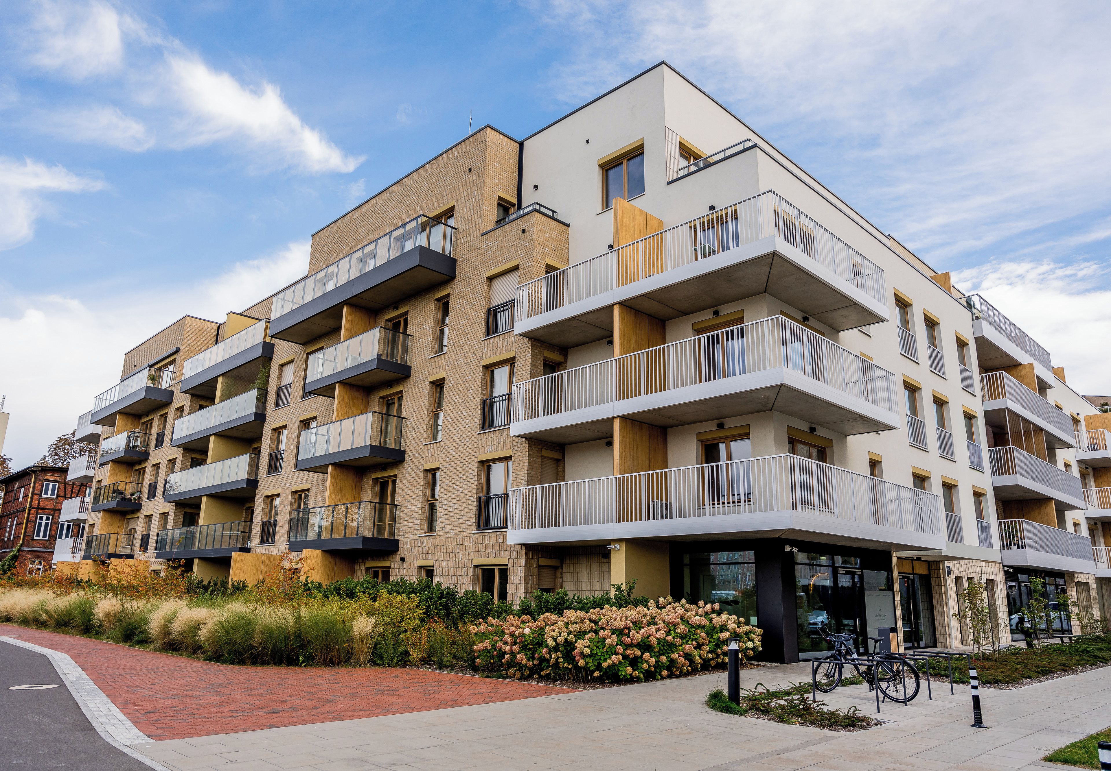A multi-story residential building with balconies, a brick facade, and a landscaped garden area.