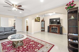 Amplia sala de estar con un sofá gris, una mesa de centro de vidrio, un tapete con estampado rojo y blanco, una chimenea, un televisor y una estantería.