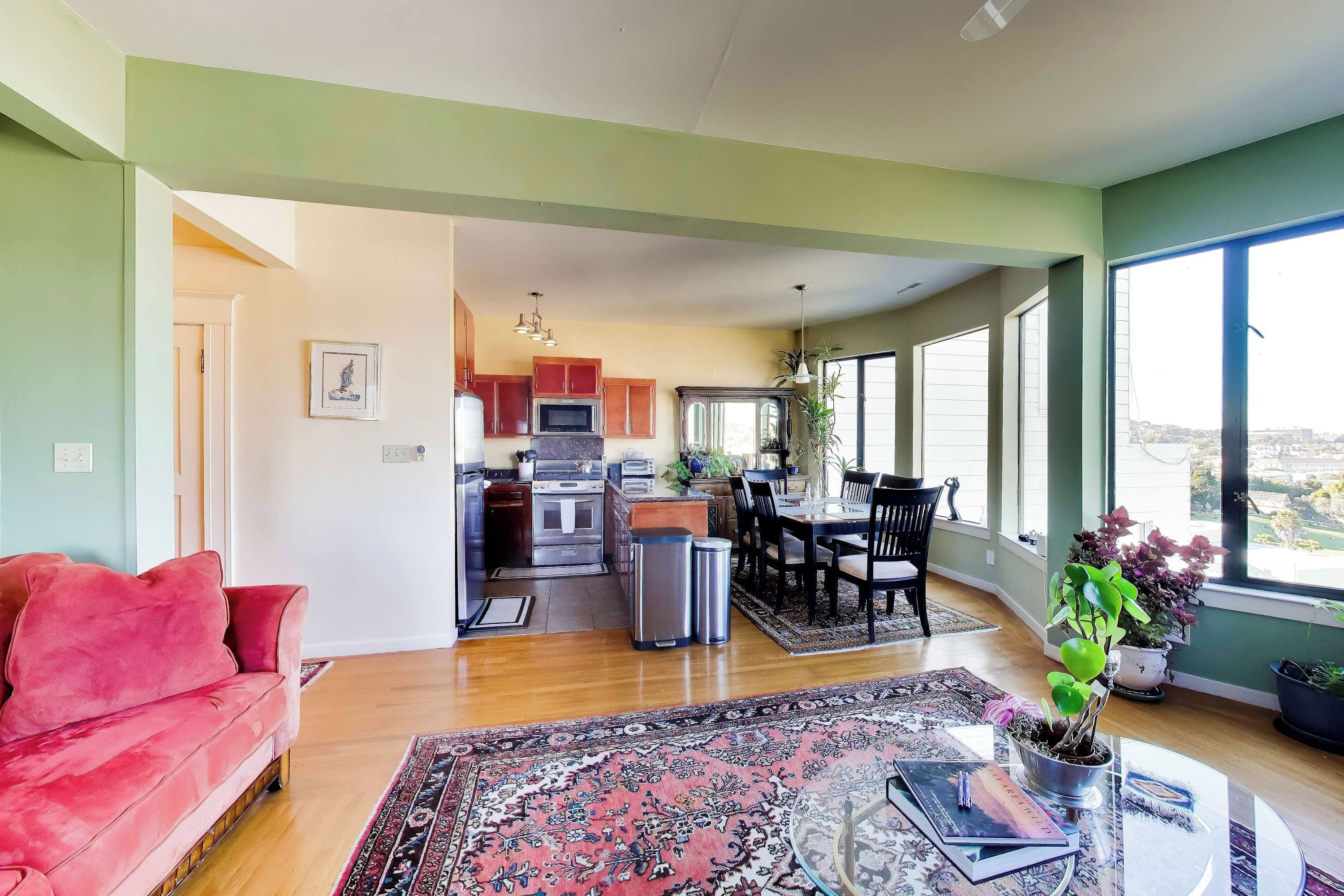 Una sala de estar moderna con un sofá rojo, un comedor con mesa y sillas, una cocina con electrodomésticos y ventanas grandes con vista a la ciudad.