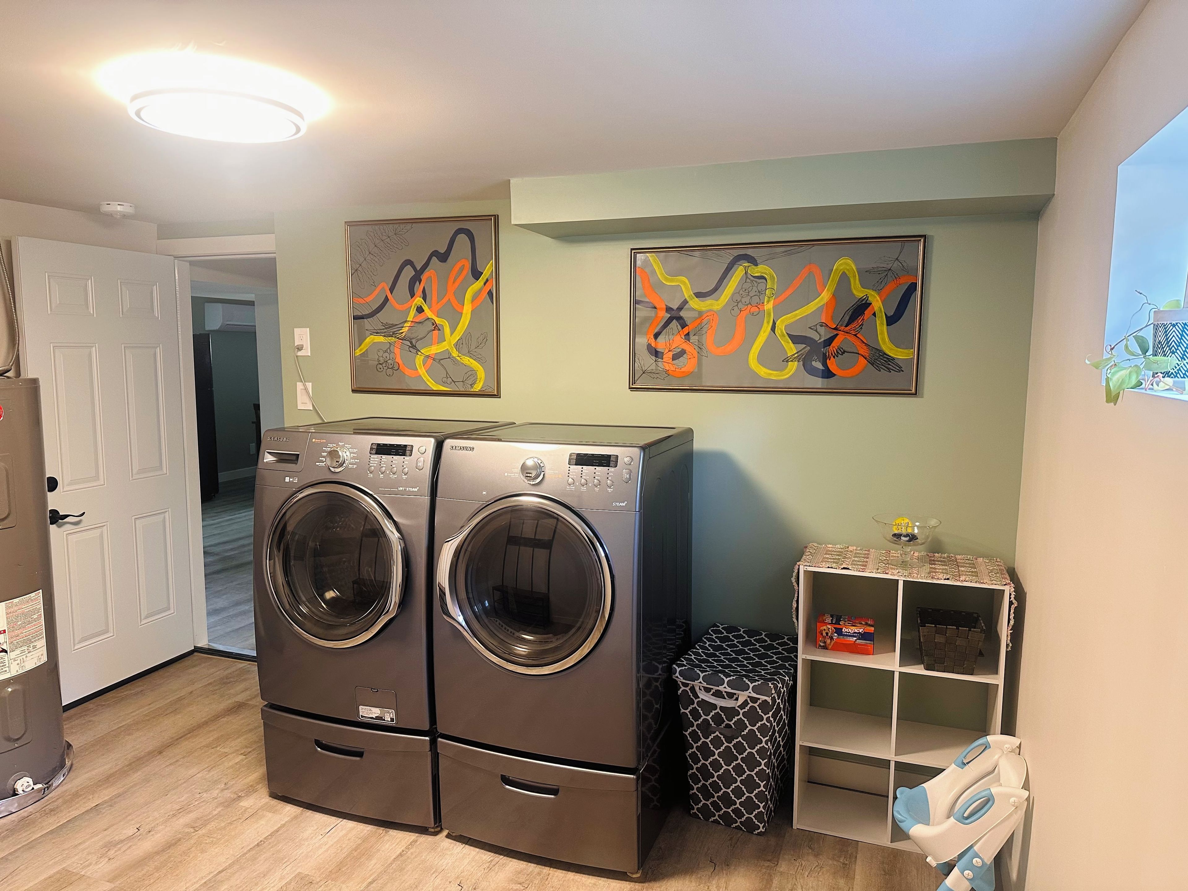 A laundry room with two stacked front-loading washers and dryers, a small shelf with a basket, and two abstract paintings on the wall.