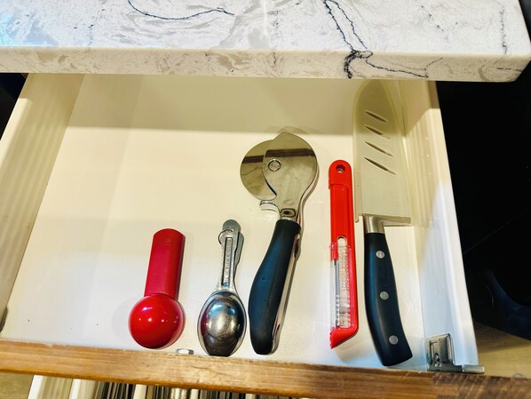 A drawer with kitchen utensils including a red-handled spoon, a silver spoon, a black-handled spatula, a red-handled grater, and a knife.
