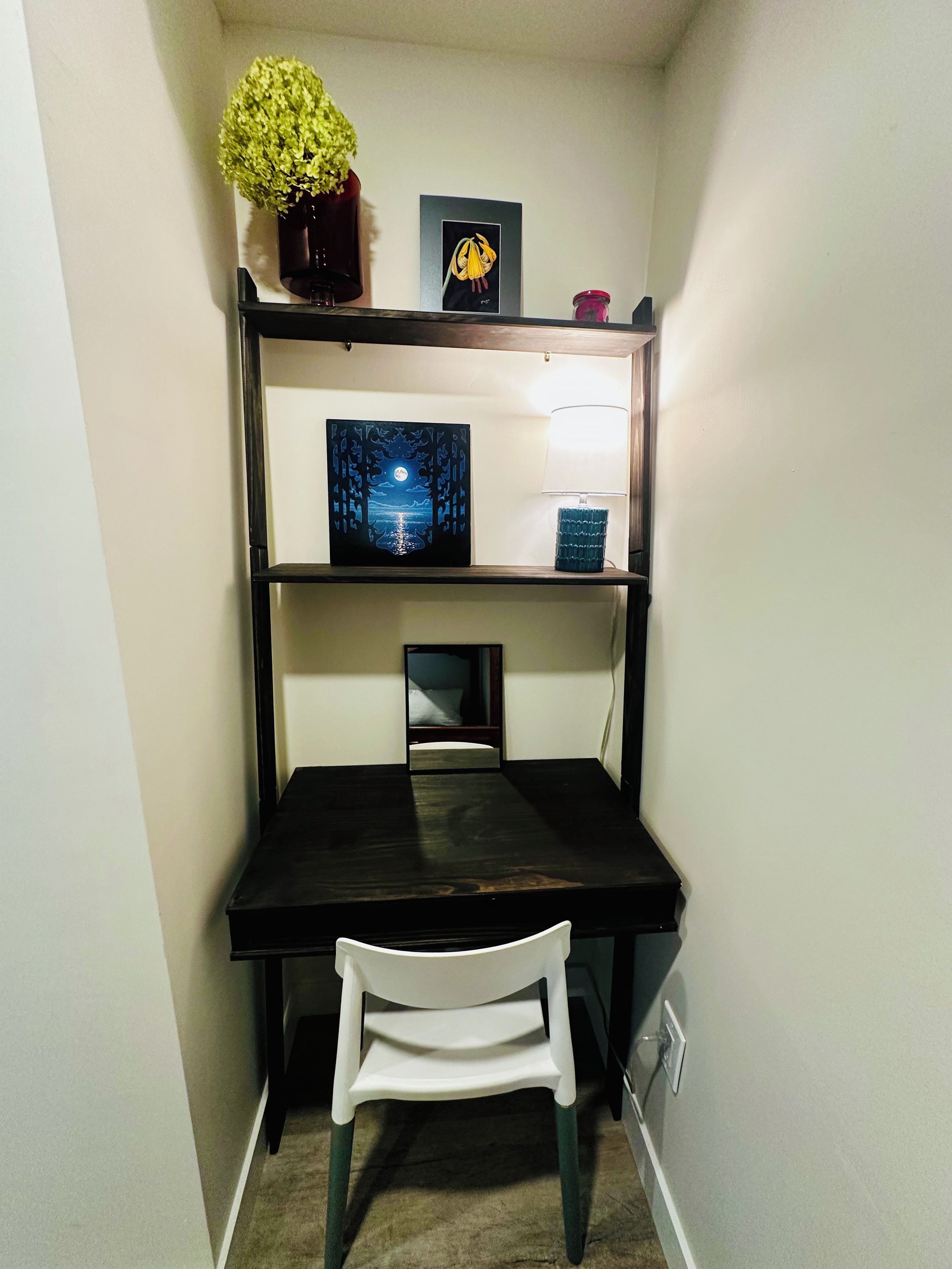 Un petit bureau avec un bureau en bois foncé, une chaise blanche, un miroir et des étagères ornées d’objets décoratifs.