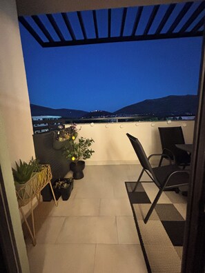 A balcony with a view of a cityscape at dusk, featuring a pergola, potted plants, and outdoor furniture.