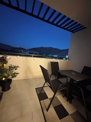 Eine Terrasse im Freien mit Tisch und Stühlen, einem Blumentopf und Blick auf eine entfernte Skyline im Abendrot.