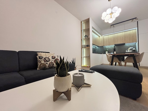 A modern living room with a dark sofa, a white coffee table with a plant and remote controls, and a kitchen in the background with a wooden cabinet and a dining area.