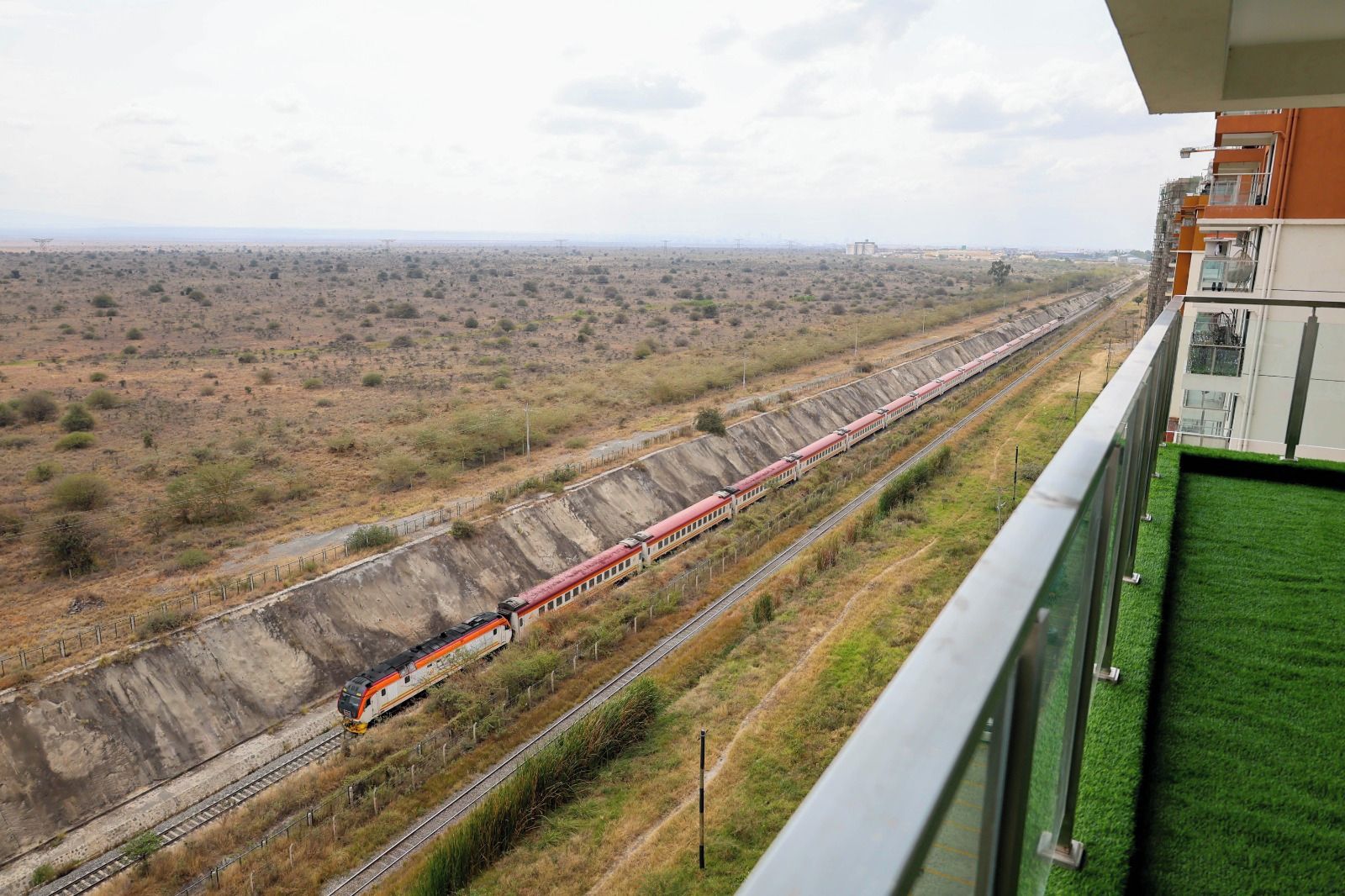 A train on tracks with a view of a barren landscape and a building with balconies.
