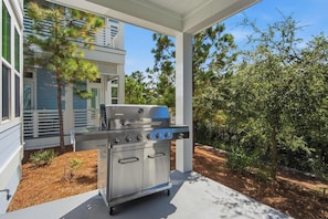 Ein Grill aus Edelstahl mit mehreren Brennern, platziert auf einer Beton-Terrasse unter einer weißen Pergola, umgeben von Bäumen und einem Haus mit blauen Fassadenplatten.