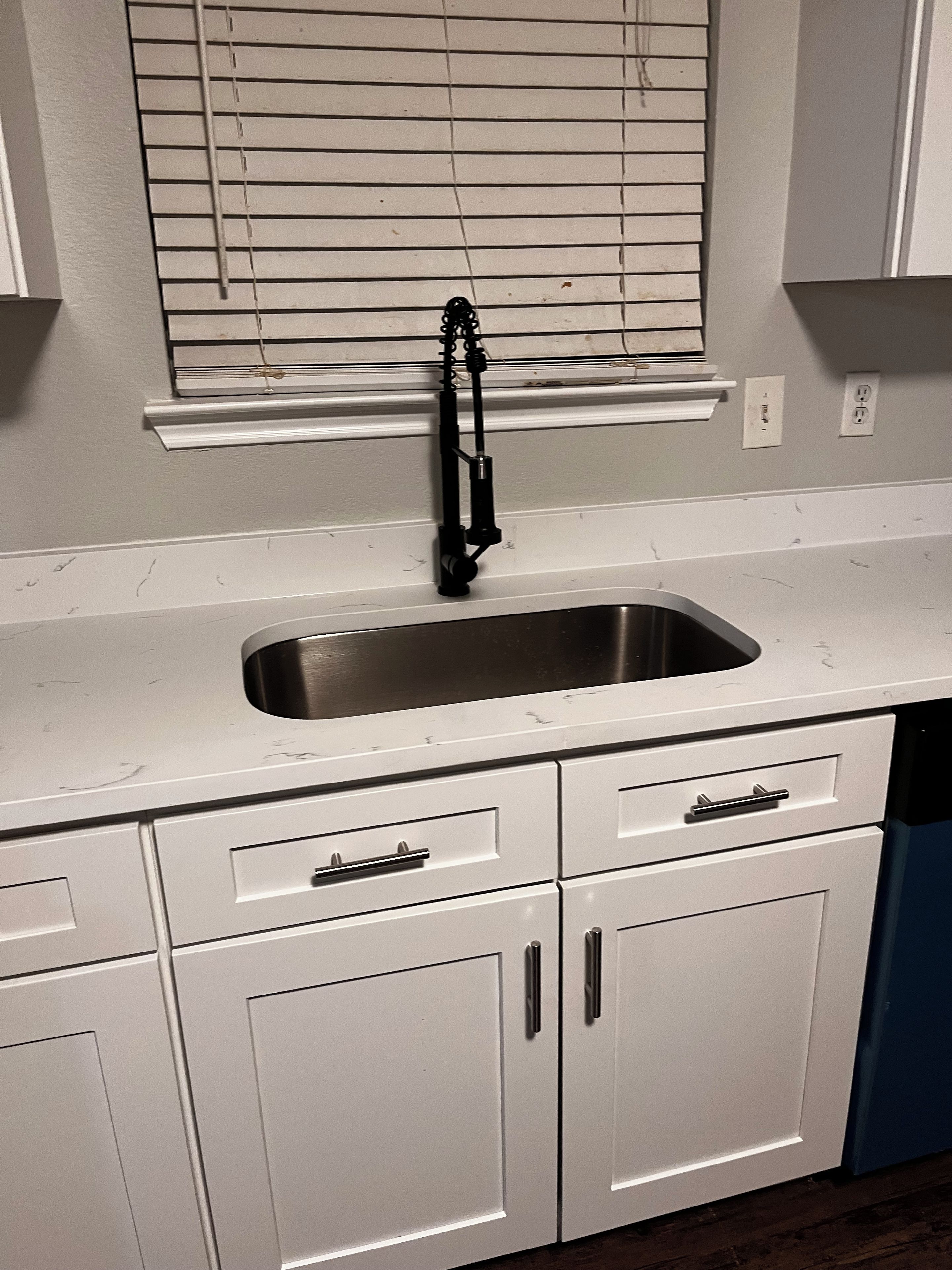 A kitchen with a stainless steel sink, white cabinets, and a window with blinds.