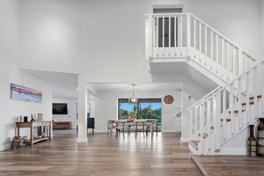 A spacious living area with a wooden floor, a dining table with chairs, a staircase with white railings, and a large window with a view of greenery. - Canalfront Pool Home near Port Charlotte • Gulf Access & Fishing Ramp (Port Charlotte)