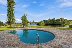 A swimming pool with a curved edge, surrounded by a brick patio, and overlooking a body of water with trees and a clear sky. - Canalfront Pool Home near Port Charlotte • Gulf Access & Fishing Ramp (Port Charlotte)