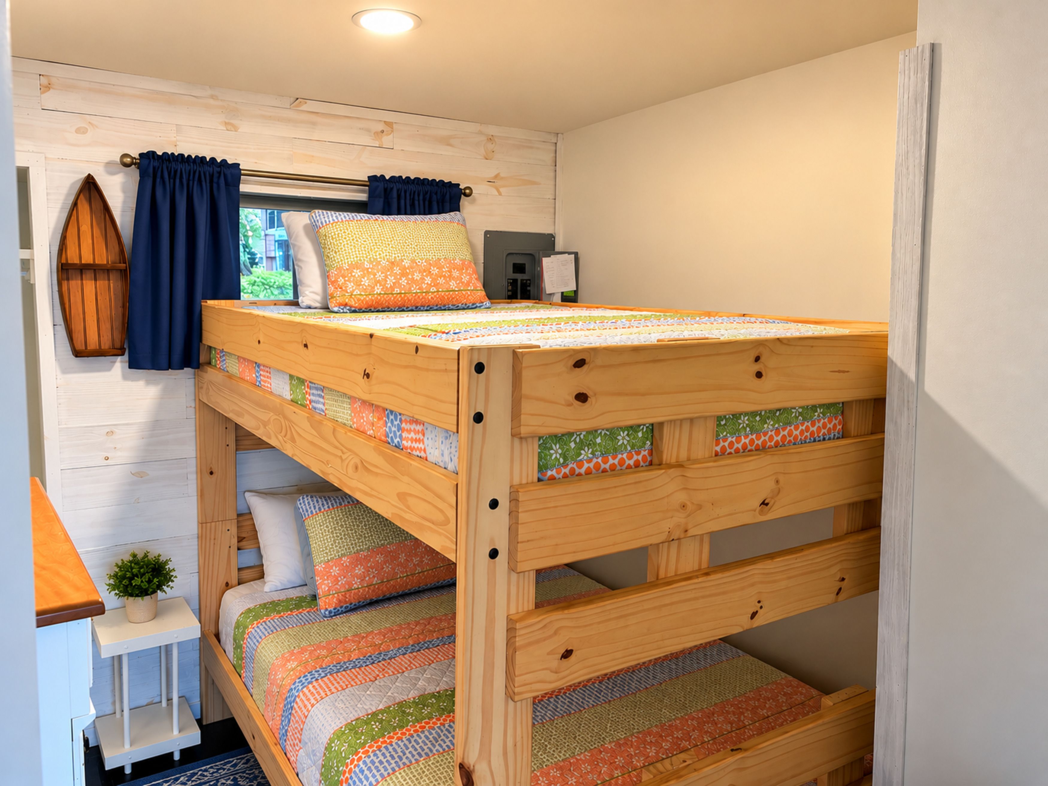 Ein Stockbett mit Holzrahmen und bunter Bettwäsche, ein kleiner Nachttisch mit Pflanze und ein Fenster mit Vorhängen.