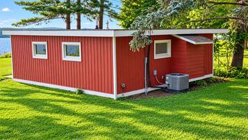 Une cabane rouge avec deux fenêtres, une cheminée et un appareil de climatisation, située sur une zone herbeuse près d’une étendue d’eau.