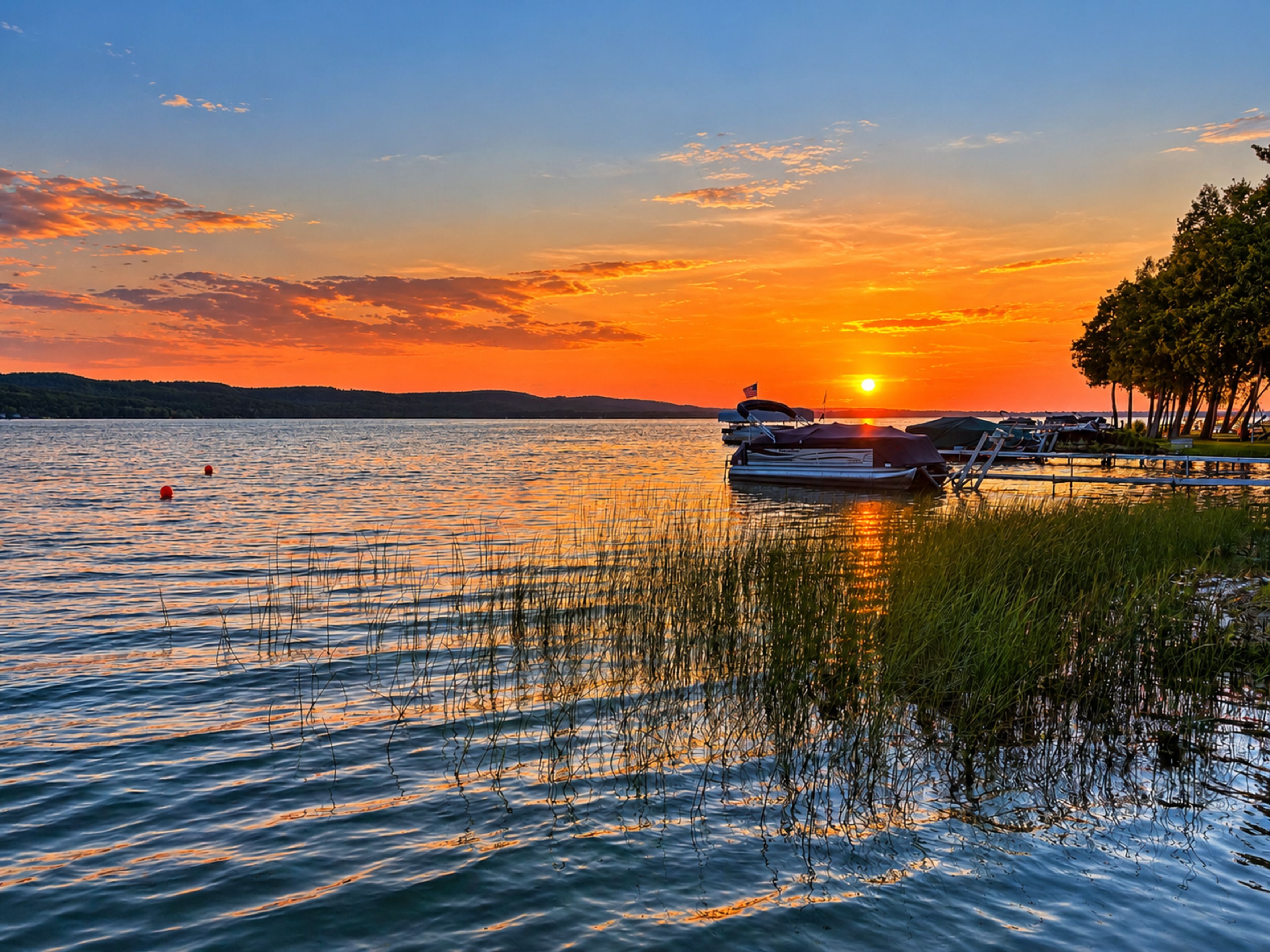 Ein ruhiger See bei Sonnenuntergang mit anlegenden Booten, umgeben von Bäumen und einem klaren Himmel.