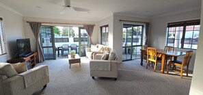 A spacious living room with a sofa, a coffee table, a TV, and a dining area with chairs and a table. - OCEAN BREEZE ESCAPE
Four Minute Walk To Beach, Town & Clubs (Whangamatā)