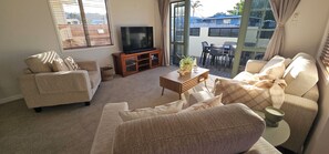 Ein Wohnzimmer mit einer Couch, einem Sessel und einem Fernseher auf einer Holzkommode. Ein kleiner Tisch mit einer Pflanze und ein Fenster mit Jalousien sind ebenfalls vorhanden.
