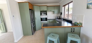 A kitchen with a refrigerator, oven, microwave, and a window with blinds. - OCEAN BREEZE ESCAPE
Four Minute Walk To Beach, Town & Clubs (Whangamatā)