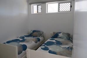 Une chambre avec deux lits simples, chacun recouvert d’une couvre-lit à motifs, une fenêtre munie d’un moustiquaire métallique et une porte blanche.