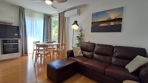 A living room with a brown leather sofa, a dining table with chairs, a standing lamp, and a painting of a sunset over water. - St Cath's Anchorage 2 Bedroom (Wynnum)