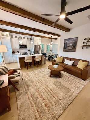 A modern living room with a brown leather sofa, a coffee table, and a dining area with a white table and chairs. - Peak on First I Modern Bungalow Near Downtown Apex (Apex)