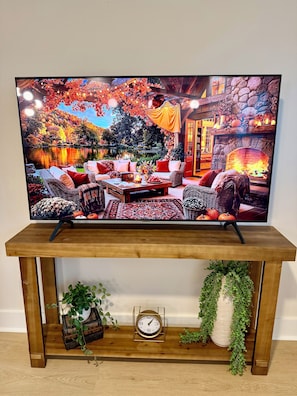 A television displaying a scenic outdoor view with a cozy seating area, a fireplace, and a wooden console table with a clock and plants. - Peak on First I Modern Bungalow Near Downtown Apex (Apex)