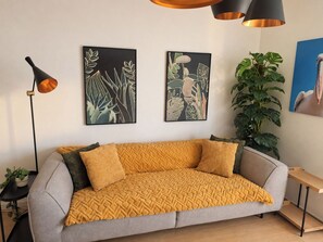 Ein modernes Wohnzimmer mit einem grauen Sofa, einer senffarbenen Decke und zwei gerahmten Pflanzenbildern an der Wand.