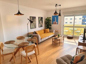 A modern living room with a glass dining table, a sofa with a yellow throw, and a large window with a view of buildings. - Lisbon • Spacious 2BR • Balcony • Elevator • WiFi (Lisboa)