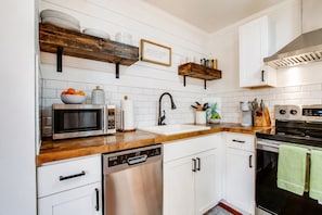 A modern kitchen with white cabinetry, a stainless steel dishwasher, a microwave, and a stove. There are wooden shelves above the counter and a framed quote on the wall. - Costy Stay | 8 minutes to Downtown & Broadway | Free Parking (Nashville)