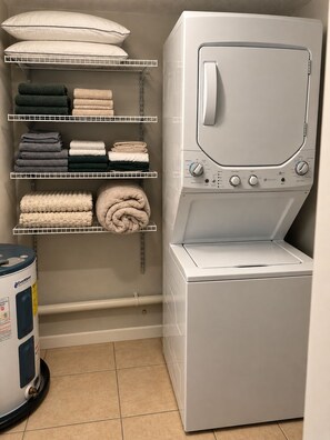 A laundry room with a front-loading washing machine, shelves with folded towels and blankets, and a water heater. - The Haus by Lioré 1BD/1BR Near Bucs Stadium & DT (Tampa)