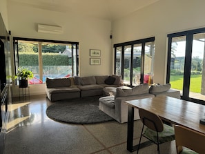 A modern living room with a sectional sofa, a dining table, and large windows offering a view of a garden. - Country resort -  Have fun with your extended family at this stylish home (Whakamārama)