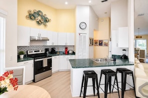 A modern kitchen with white cabinets, a central island with bar stools, a clock on the wall, and a decorative wall piece. - Lakefront Pool & Theater Near Disney (Kissimmee)