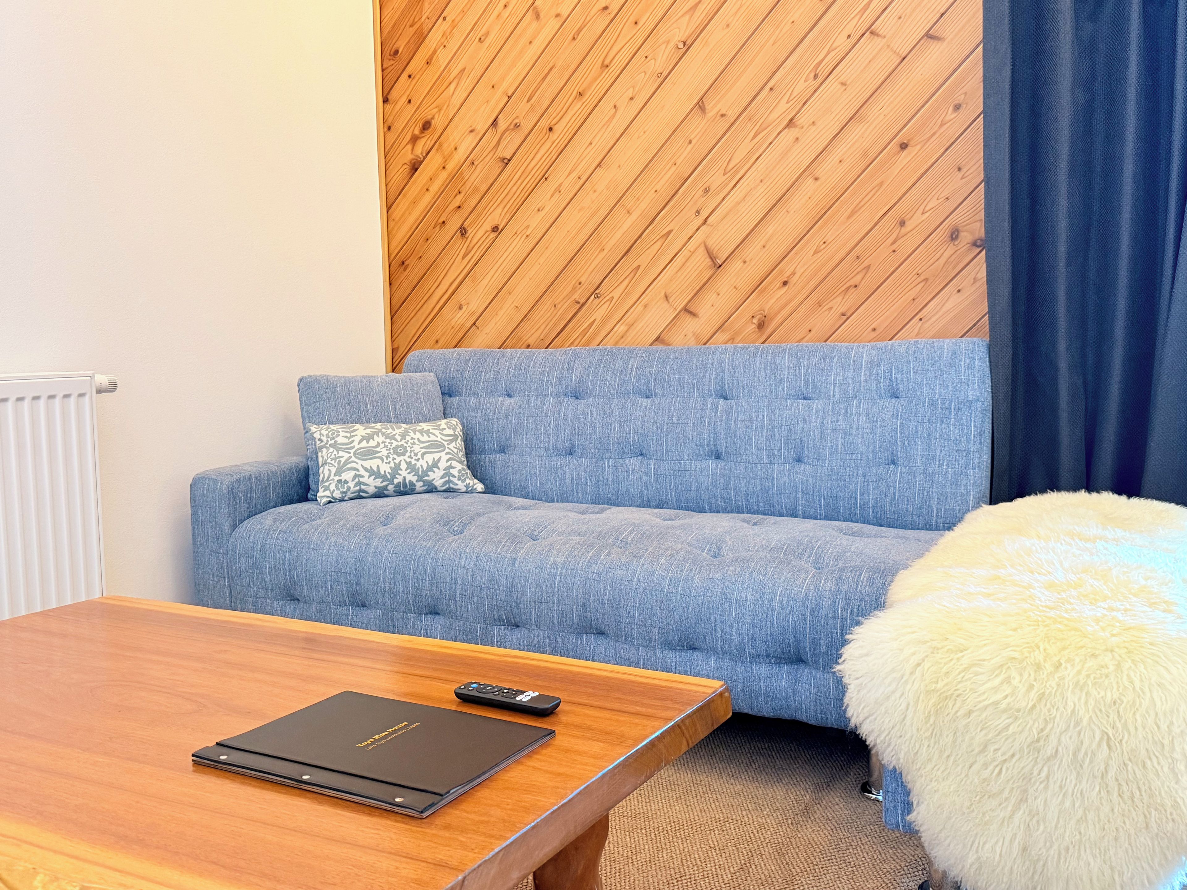 A room with a blue sofa, a wooden coffee table, a remote control, and a fluffy white ottoman.