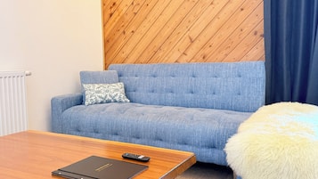 A room with a blue sofa, a wooden coffee table, a remote control, and a fluffy white ottoman.
