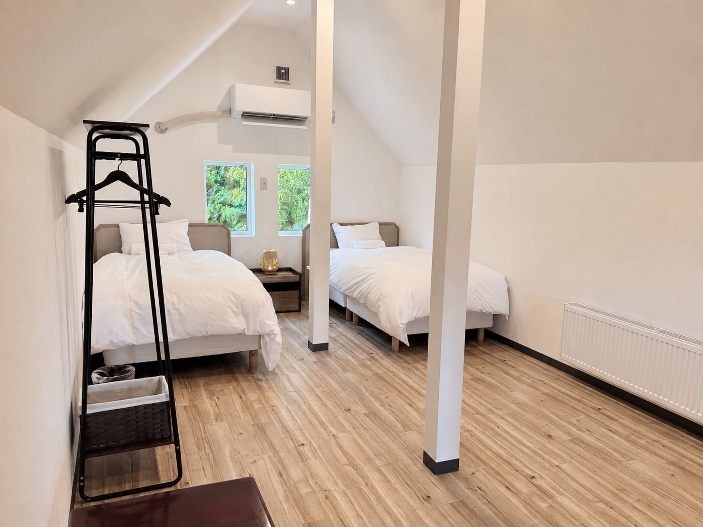 A hotel room with two beds, a wooden floor, a hanging chair, and a window with a view of greenery.
