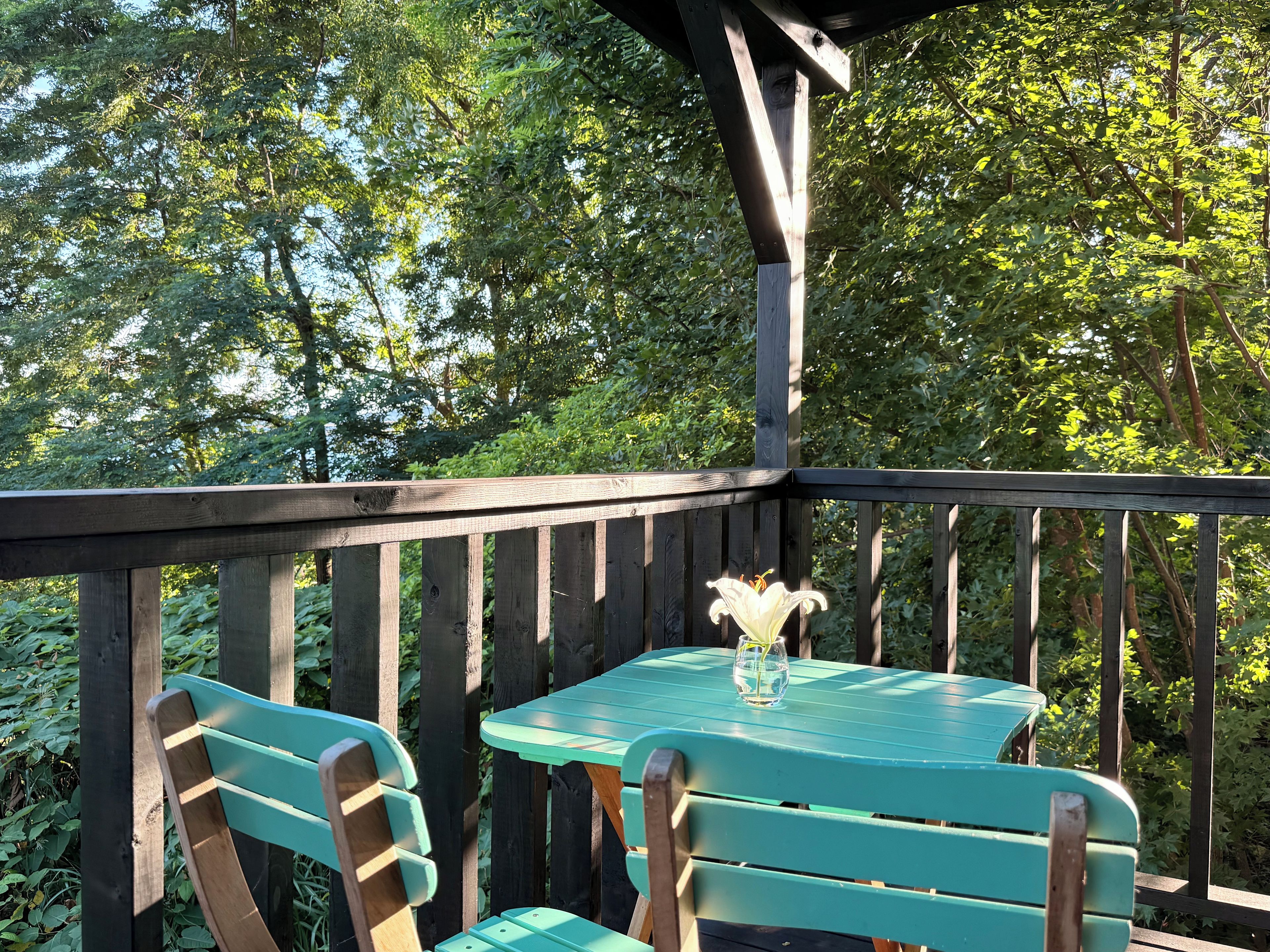 A wooden deck with two green tables and chairs, surrounded by lush greenery.