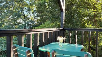 A wooden deck with two green tables and chairs, surrounded by lush greenery.