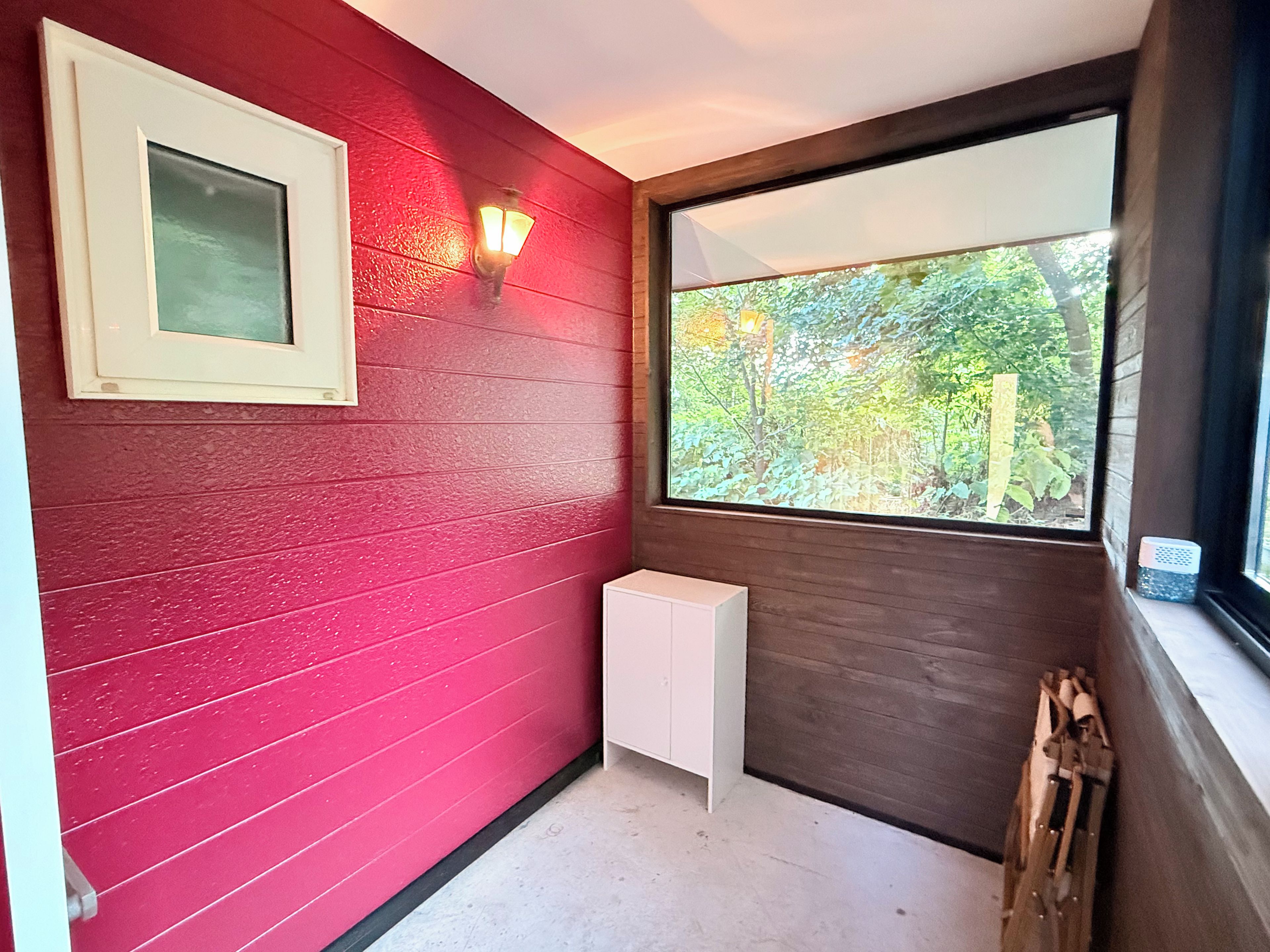 A room with a red wall, a white cabinet, a window with a view of greenery, and a wall-mounted light fixture.