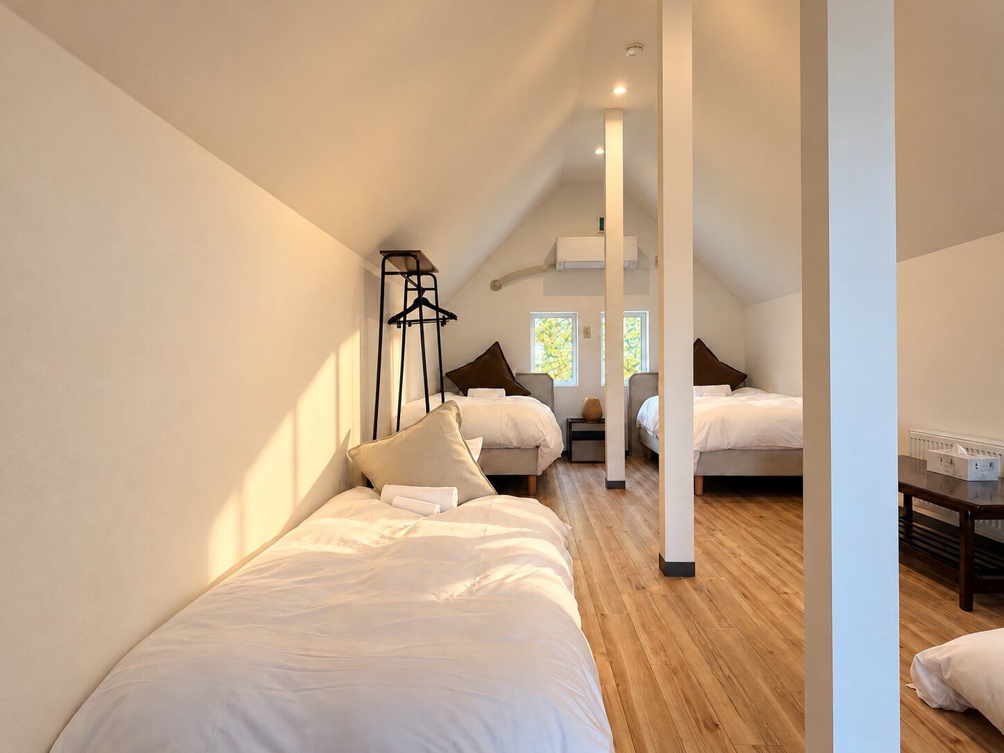 Ein modernes Hotelzimmer mit schrägem Dach, Holzboden und einer zentralen Säule. Es gibt zwei Betten mit weißen Bettwäsche und einen kleinen Tisch mit einer Lampe.