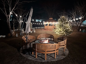 Eine Terrasse im Freien mit Holzstühlen und einem Feuerkorb, umgeben von Bäumen und einem Pavillon mit Lichterketten.