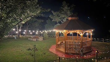 Un jardín con un quiosco de madera y base de piedra, rodeado de un sendero rojo y luces colgantes.