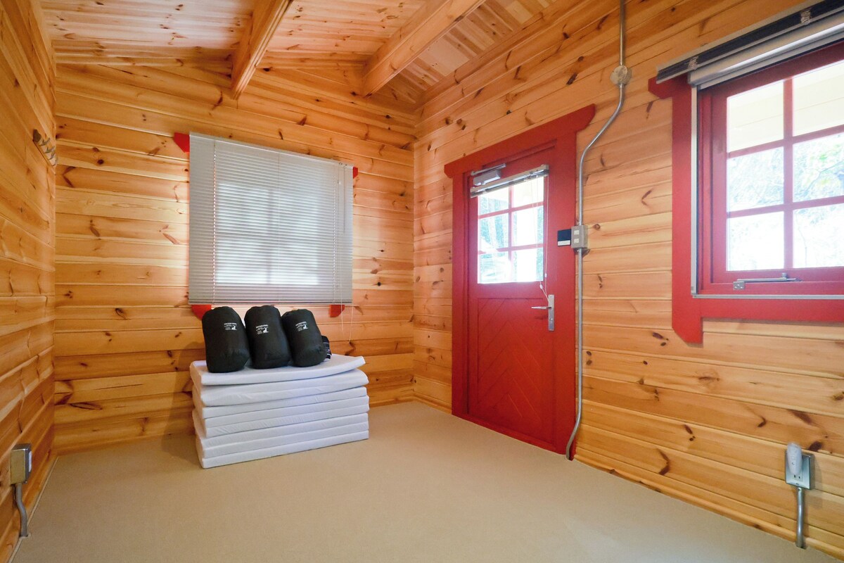 Interior de una cabaña de madera con una puerta roja, una ventana con persianas y una pila de colchones blancos con almohadas negras.
