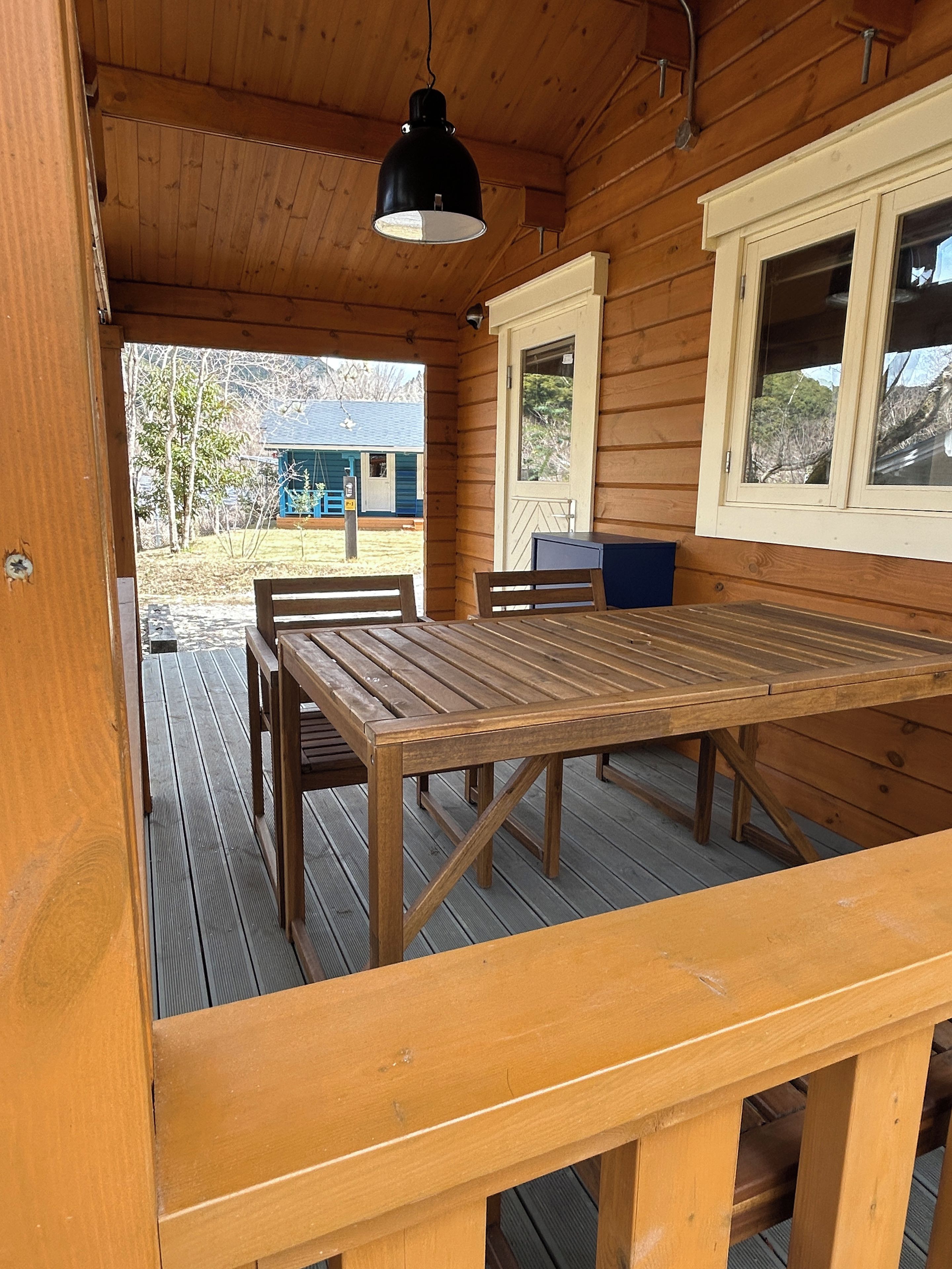 Una terraza de madera con una mesa, sillas, una lámpara colgante y ventanas con vistas al exterior.