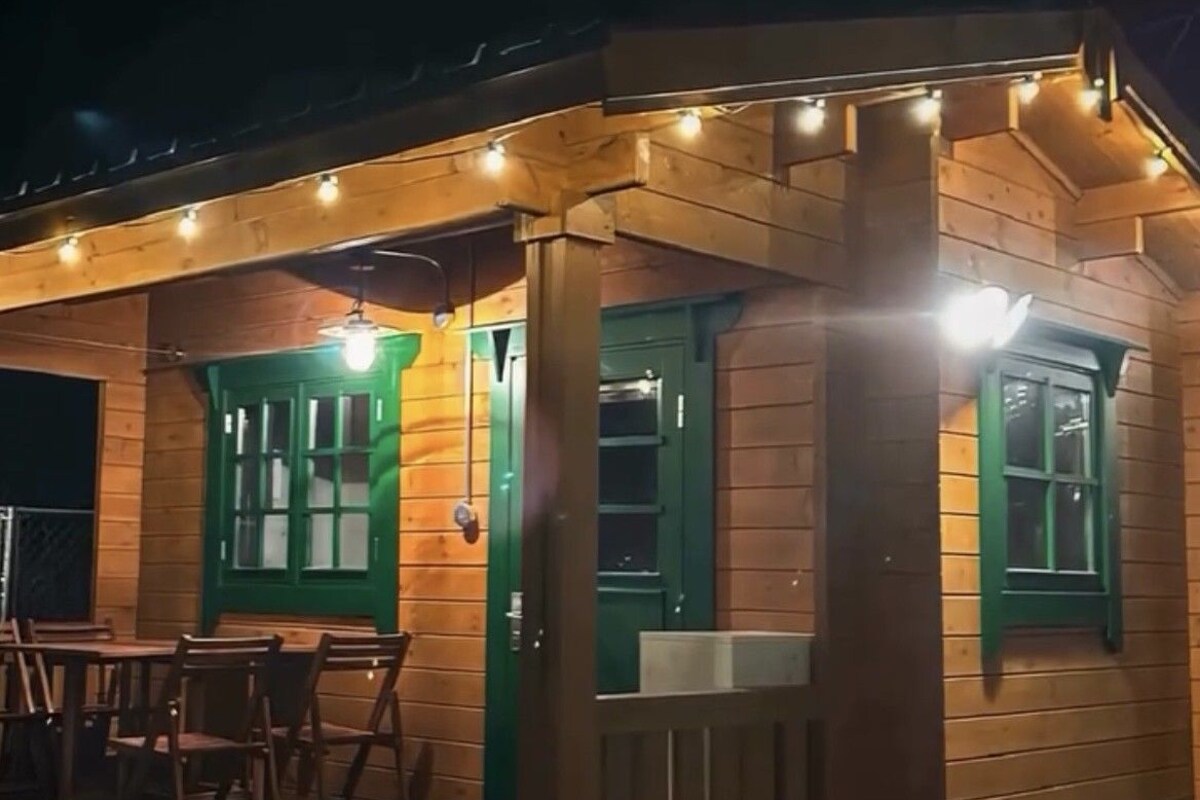 A wooden building with a covered porch, green-framed windows, and a seating area with tables and chairs.