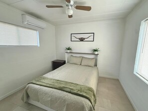 Une chambre à coucher comprenant un lit, un ventilateur de plafond, un appareil de climatisation, une table de chevet, un tableau encadré au mur et une fenêtre avec des stores.