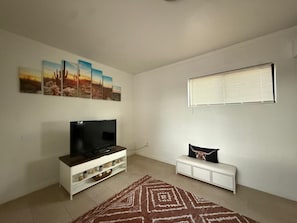 Ein Wohnzimmer mit einem Flachbildfernseher auf einer weißen Unterhaltungseinheit, einem gemusterten Teppich, einem weißen Beistelltisch mit einem dekorativen Kissen und einem Fenster mit Jalousien.