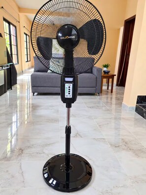 A standing fan with a control panel, placed on a tiled floor in a hallway with a sofa and a small table in the background. - Casa del Marino (Tela)