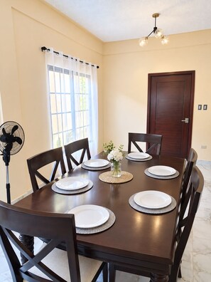 A dining room with a wooden table set for six, chairs, a fan, a door, and a window with curtains. - Casa del Marino (Tela)
