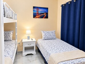 A bedroom with two beds, a nightstand with a lamp, a painting of a bridge, and a blue curtain. - Casa del Marino (Tela)