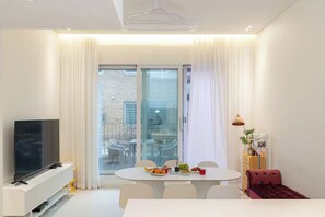A modern living room with a flat-screen TV, a dining table with chairs, a red sofa, and a large window with curtains.